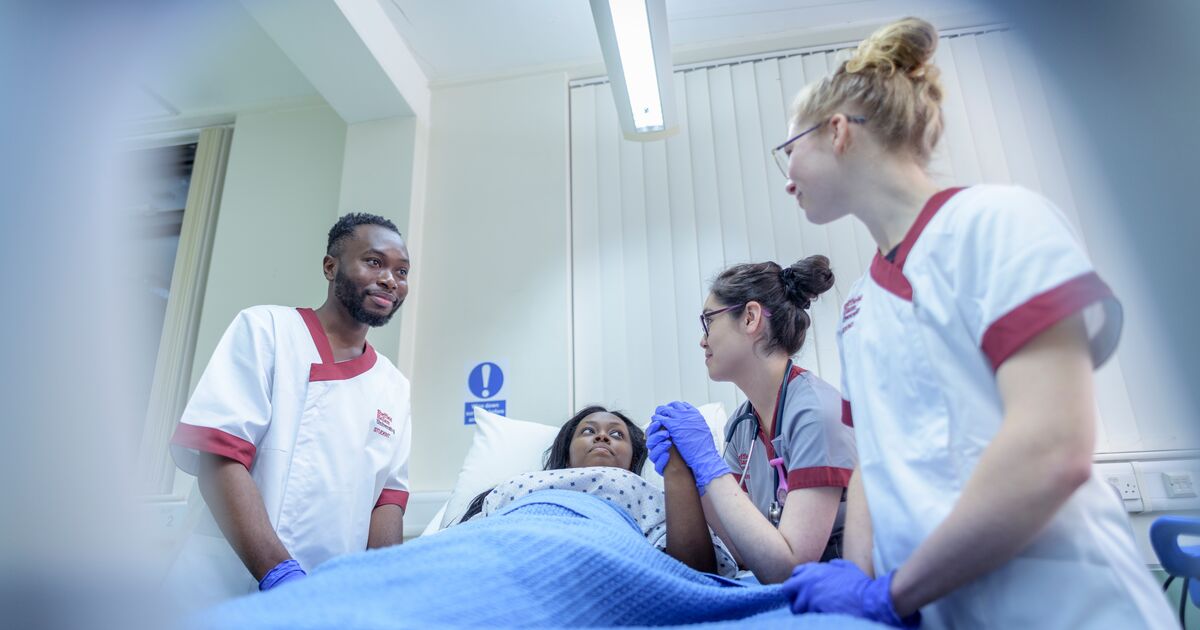 Image shows three student nurses gathered around a hospital bed, which has a patient in it. One of the student nurses is holding the hand of the patient.