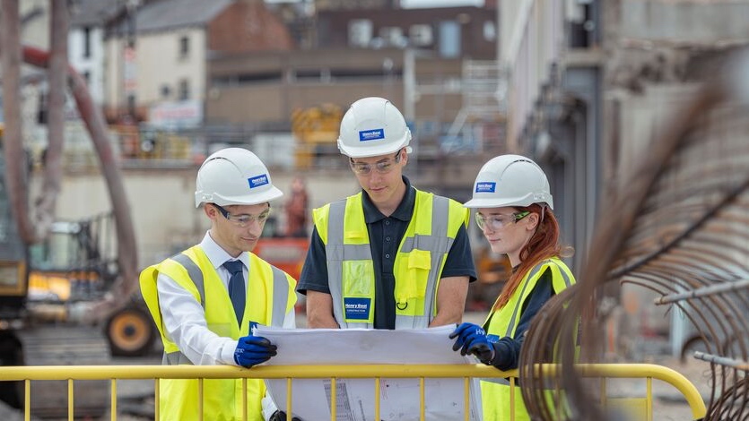 Three apprentices wearing hi-vis jackets and hard hats stood on a construction site looking at a printed plan