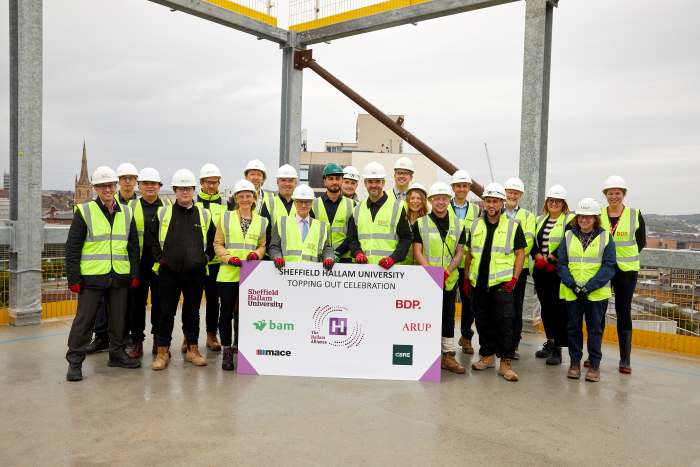 Image shows a group photo of Hallam Alliance on top of one of the new buildings on Howard Street. They are standing behind a big sign with Hallam Alliance logos on. All are wearing hi-viz and helmets