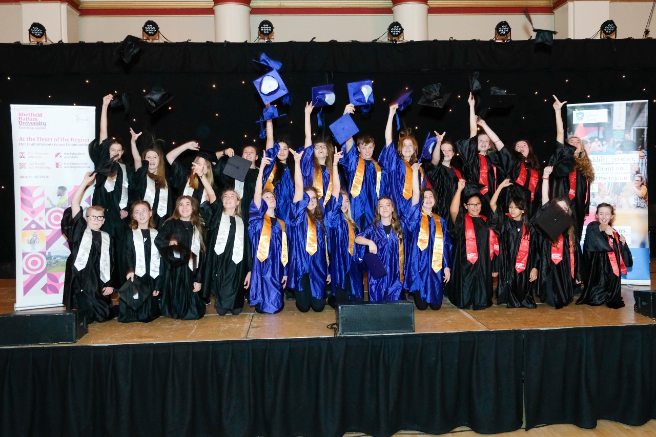 Children are on a stage dressed in graduation caps and gowns, as a group they are throwing their graduation caps in the air.