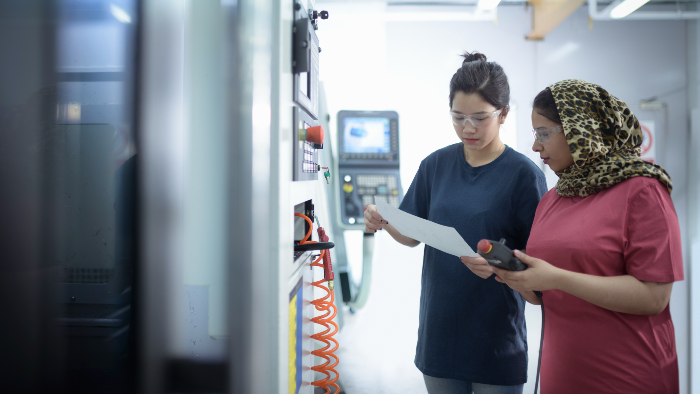 A student and academic examine a piece of paper, they are standing in front of machinery 