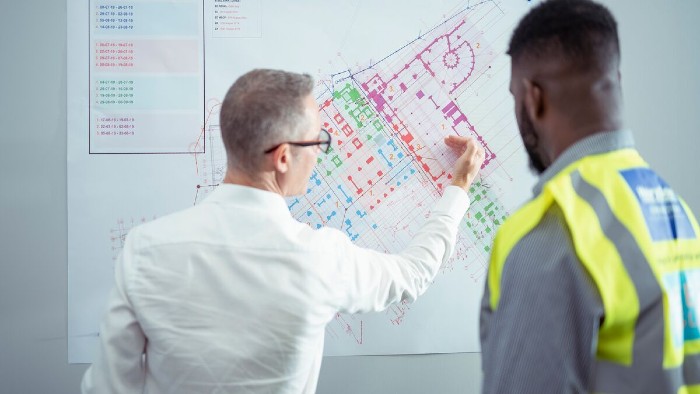 A businessman and a student in a high visibility jacket review a site plan on a whiteboard