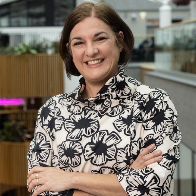 Image shows Dr Jo Lidster smiling at the camera. She has her arms folded in a relaxed way. She is wearing a white blouse with black flower patterns on.