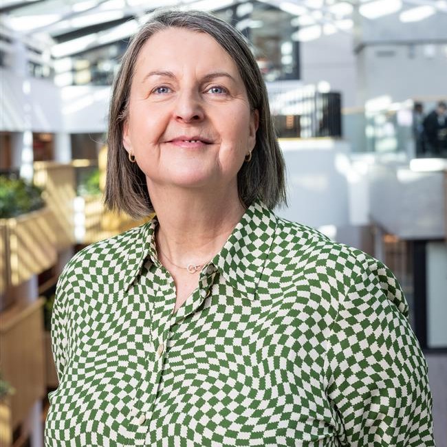 Image shows Sarah Pearson, wearing a green and white checked blouse. She is smiling at the camera, and standing in the atrium.