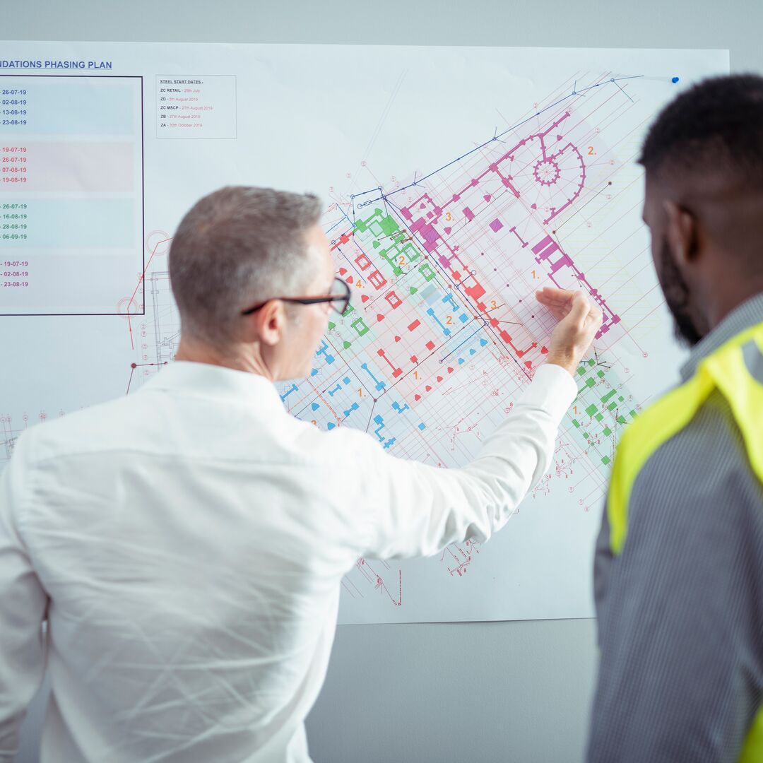 Image shows a construction blue print on a whiteboard, which a tutor is pointing to whilst a student wearing a high visibility jacket looks at the board too.