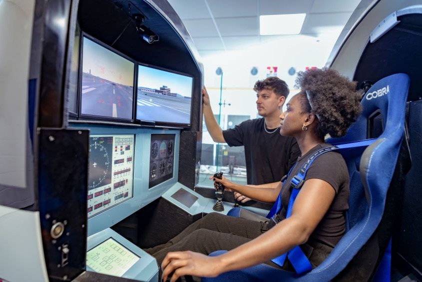 Image shows female student sitting in a driving seat. She is looking at two screens and her hand is on a gear stick. There is a male student watching the screens too.
