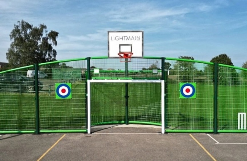 Outdoor sports court with green fencing, basketball hoop, football goal and colourful target panels.