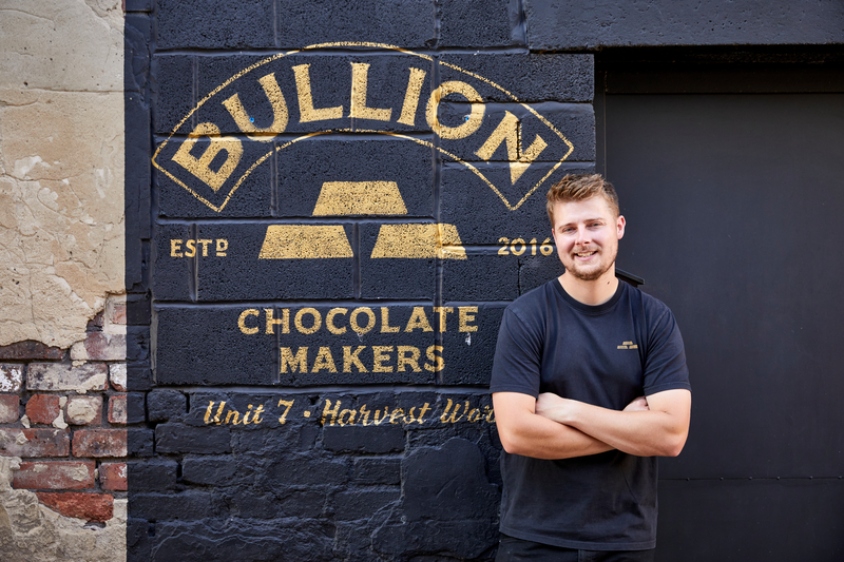 Image of Max standing in front of his business, Bullion Chocolate, which is painted on a black brick wall in gold paint