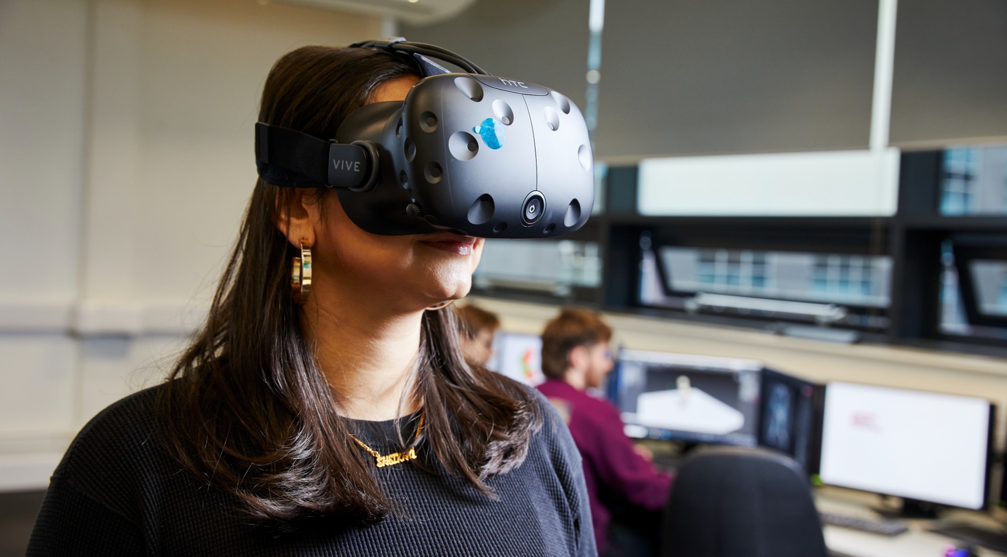 Student wearing VR headset in a classroom. In the background are two other students working at PCs.