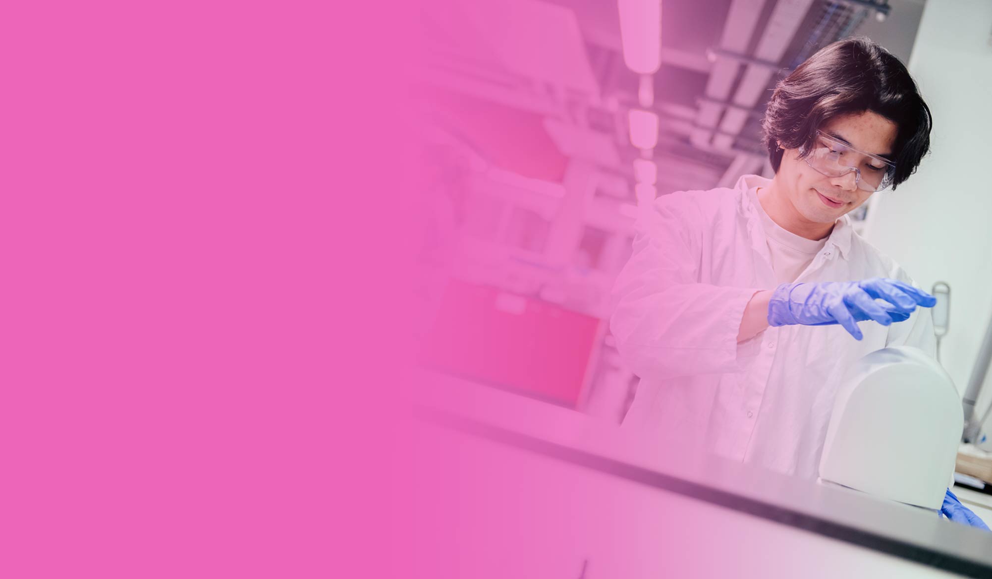 Male student in protective equipment working in a lab, against a pink gradient background