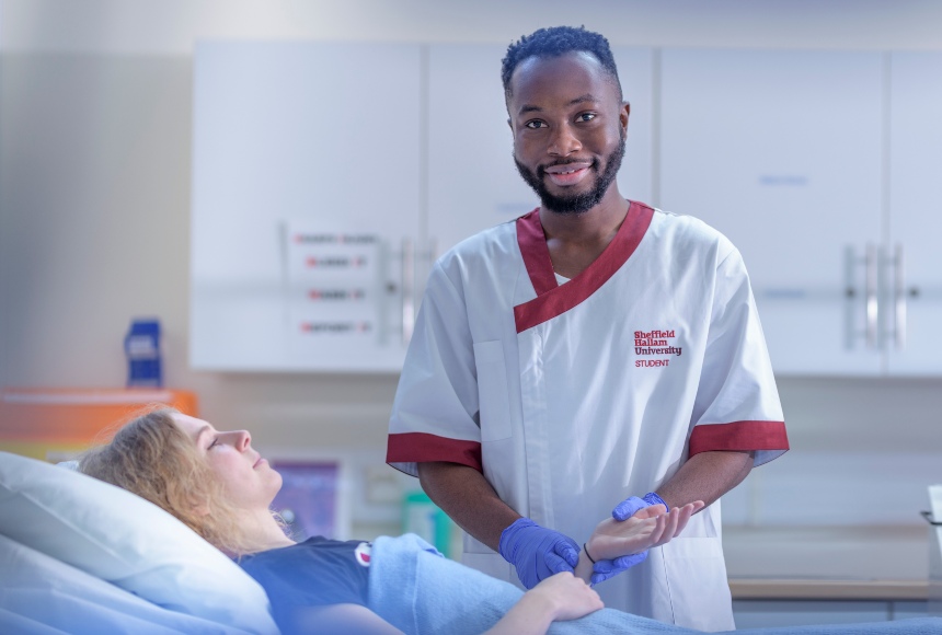 Black male nurse with facial hair smiling and standing over white blonde hair female patient in hospital ward.