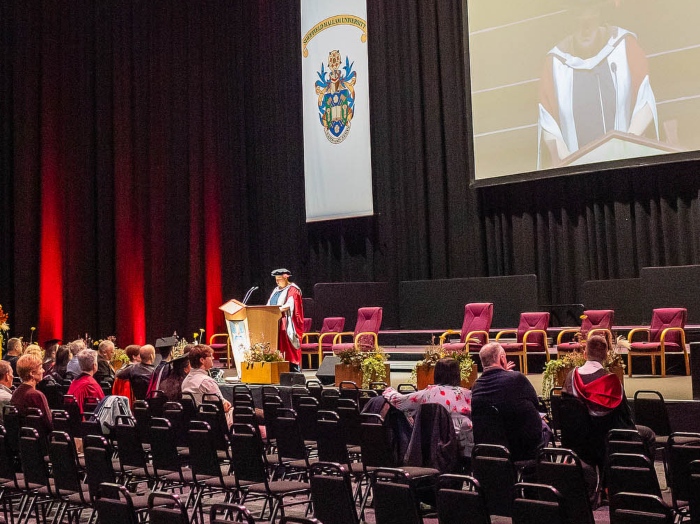 A person in academic regalia speaks at a podium on a stage with empty chairs; audience members sit in rows facing the stage at a graduation ceremony.