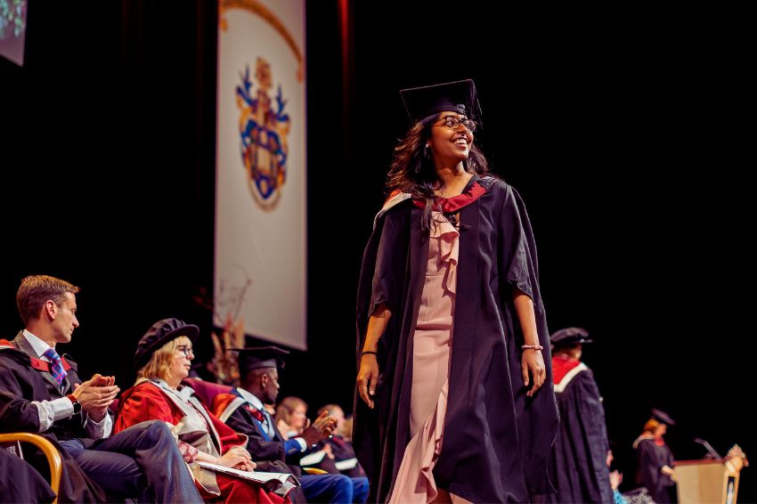 A person wearing a Graduation gown, walking across a stage during a Sheffield Hallam University Graduation ceremony