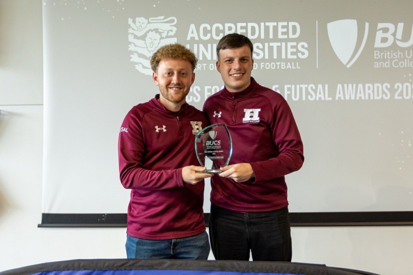 Two individuals in matching maroon jackets with an "H" logo hold a glass award and smile at the camera. Behind them is a BUCS Football & Futsal Awards 2023 banner celebrating accredited universities in English football.