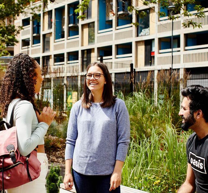 Three young people outside a block of flats in Sheffield. There are two women standing up in conversation, and a man seated next to them, listening. Immediately behind them there is a patch of grass, wildflowers and small trees.