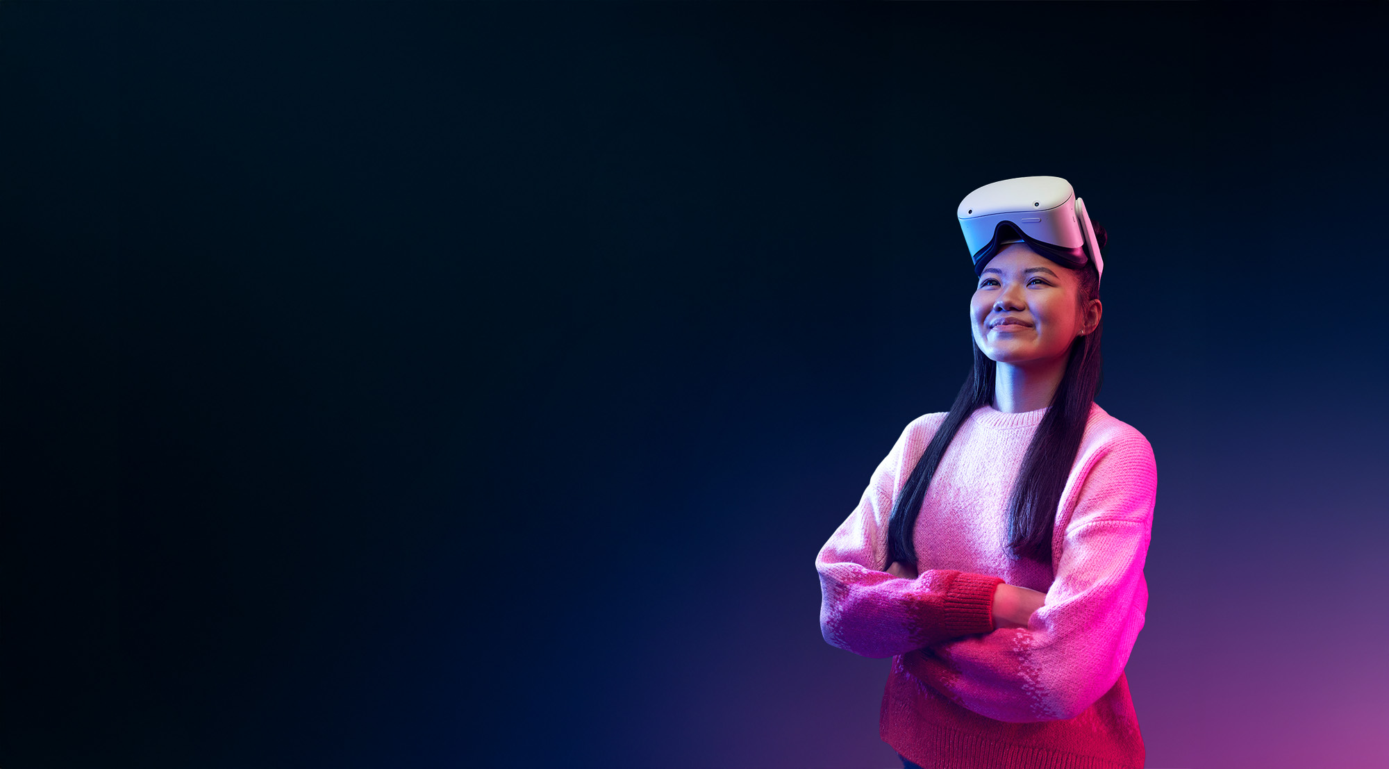 A smiling student wearing a virtual reality headset on the top of their head and a pink and white sweater, standing with arms crossed against a dark blue background.