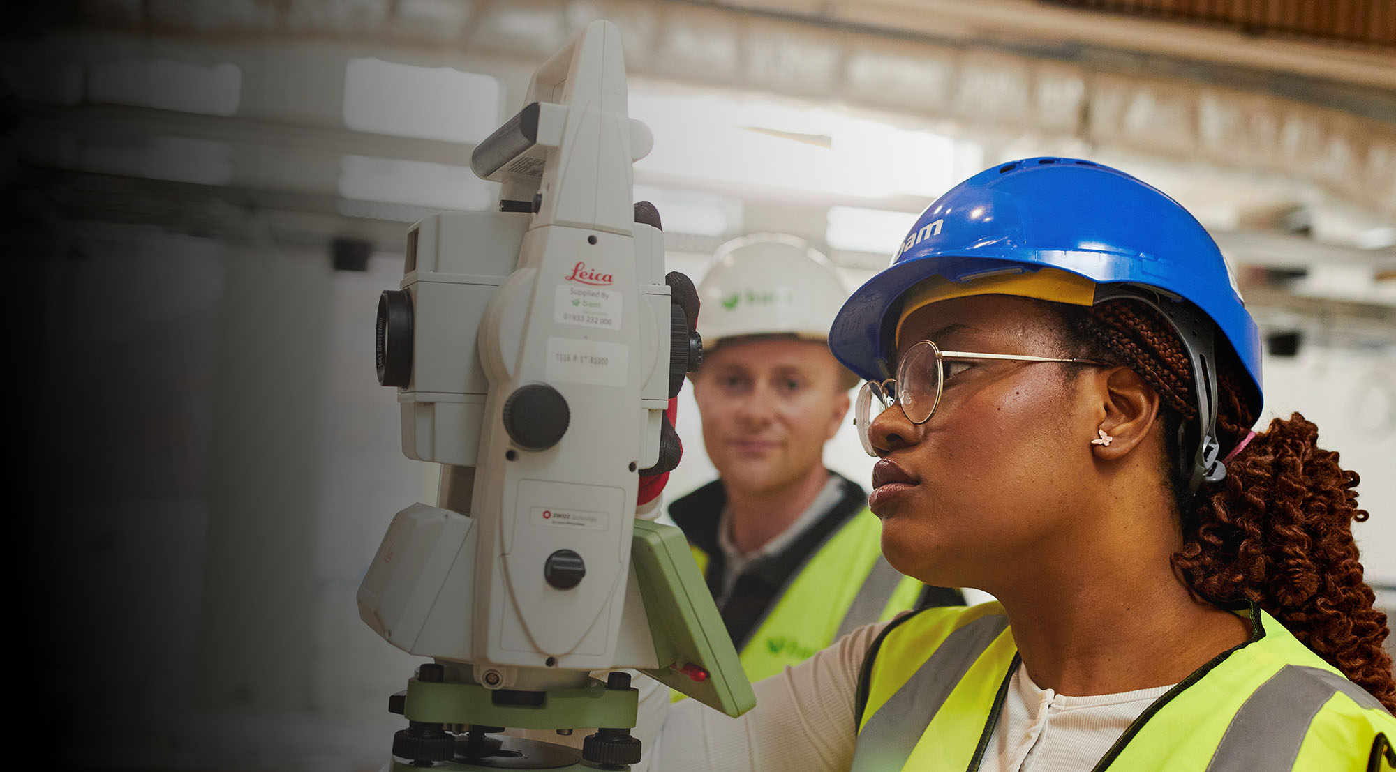 A female student wearing a blue hard hat and high-visibility vest using a surveying instrument at a construction site.