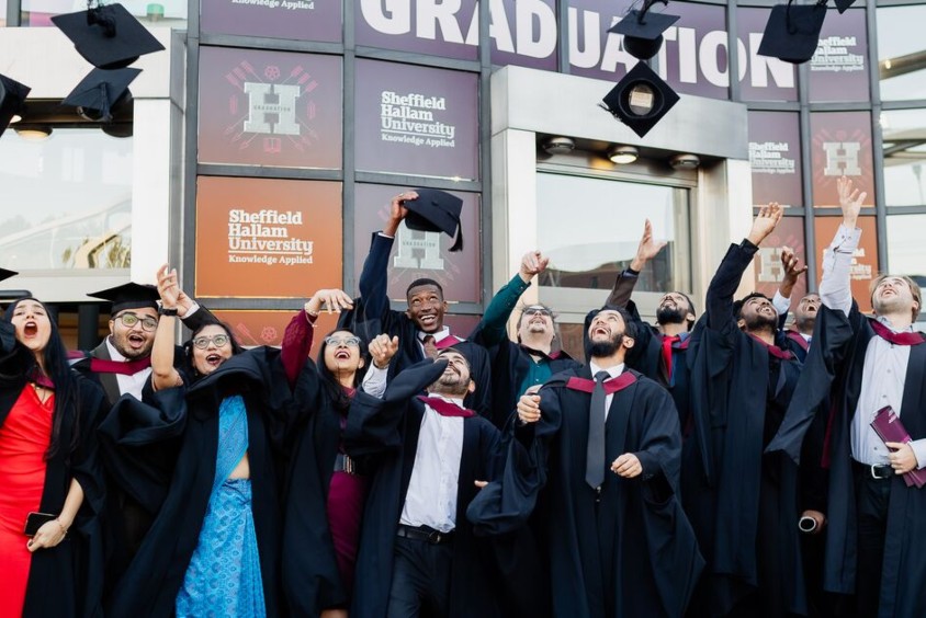 A group of Sheffield Hallam graduates wearing gowns celebrate by tossing their caps into the air in front of Ponds Forge Sports Centre.