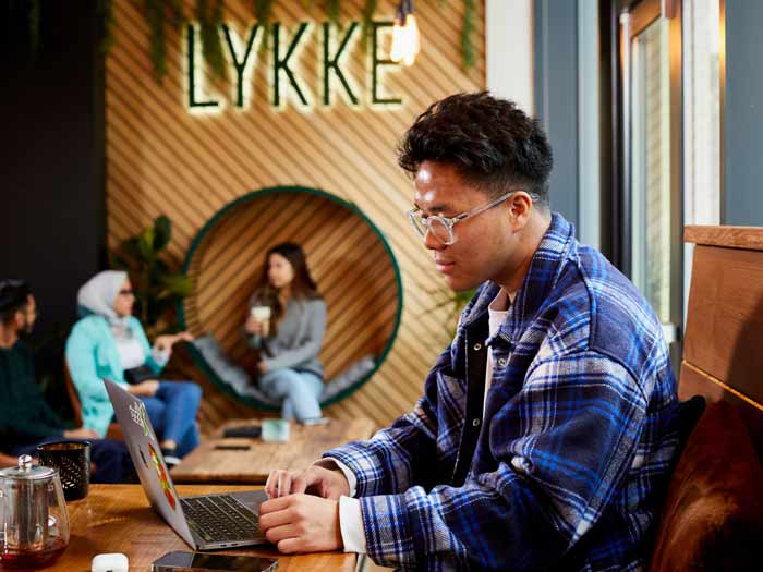 Students in a cafe in Sheffield city centre. A young man sits at a table working on his laptop. In the background we can see three students talking in a spacious room lit by hanging bulbs and floor-to-ceiling windows, with wooden chairs, benches and wall panels.