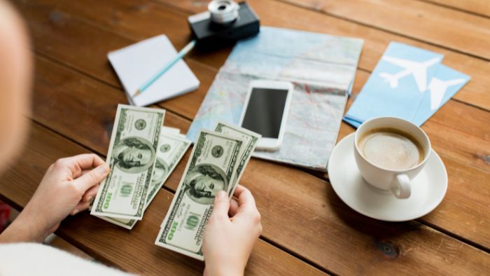 A person counting money on a table with a cup of coffee, mobile phone, digital camera, map and plane tickets.