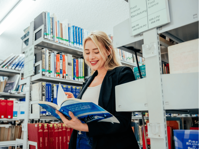 Blonde-haired student standing in library stacks, looking at the book she is holding in her arms