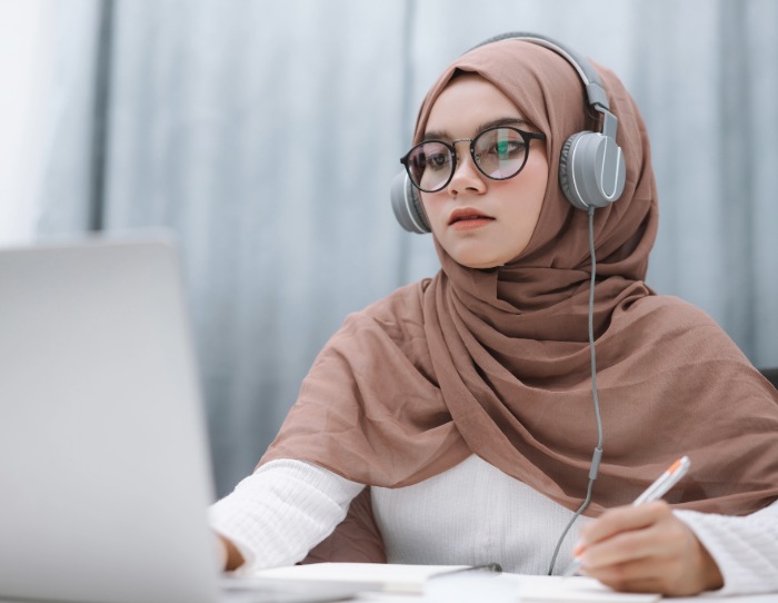 A student wearing a brown hijab and headphones takes notes while working from a laptop.
