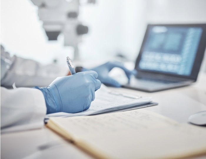 A scientist takes notes for a report with medical equipment and a laptop in the background.