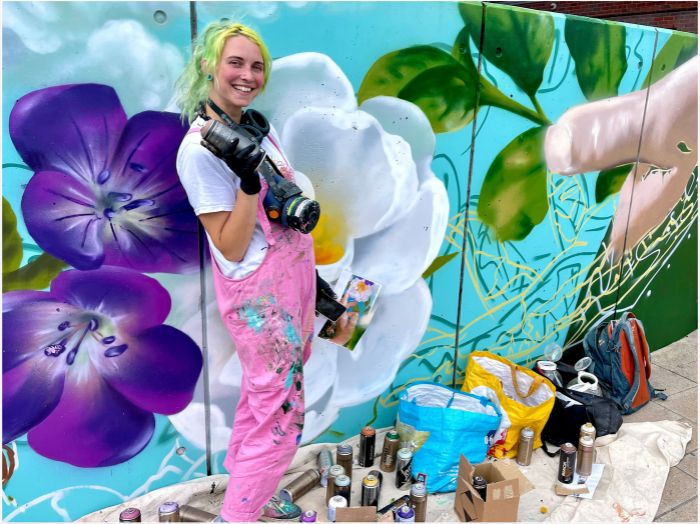 An artist standing in front of a mural she has painted, holding spray paint with flowers and bees painted in the background.