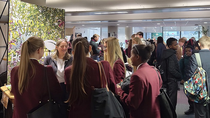 A group of children in burgundy school blazers talk to a person stood in front of a banner with trees on inside a University building