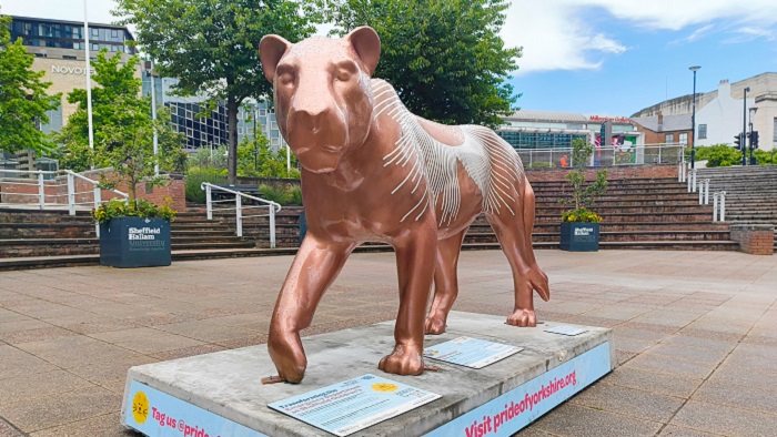 A lioness sculpture painted gold with white sound wave lines on its back. There is a paved area and trees in the background. 