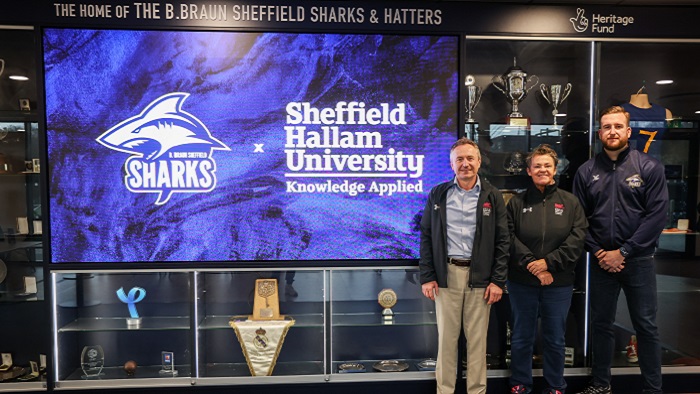 Three people stood smiling at the camera next to a trophy stand and a large screen with the logos of Sheffield Hallam University and the Sheffield Sharks
