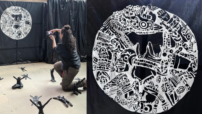 On the left, Helen Blejerman kneels to take a photograph whilst working on the art installation, around her are small figures made from black rubbish bags and tape. On the right is a painting of the Aztec Coyolxāuhqui stone.