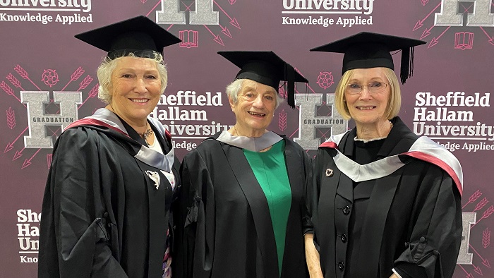 Three people wearing university cap and gowns stood in front of a maroon back drop with the words Sheffield Hallam University on it