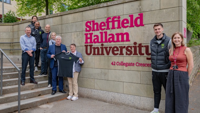Spencer Taylor from Inspire Development Academy, holds a football shirt with Dr Ruth Crabtree, Head of the School of Sport & Physical Activity, in front of a Hallam sign with more team members for the launch of a dual 'study and play' football partnership.