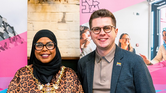 Two people stood in front of a banner with images of people on a pink background