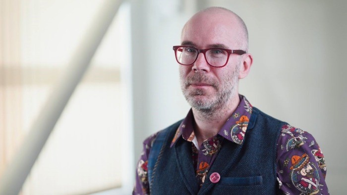 Peter Macqueen portrait photograph looking to camera. Peter is wearing red framed glasses and has a bright, patterned shirt. 