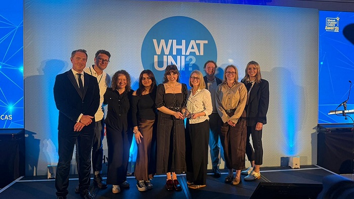 A group of people stood on a stage holding a glass trophy with a white backdrop with the word What Uni on it