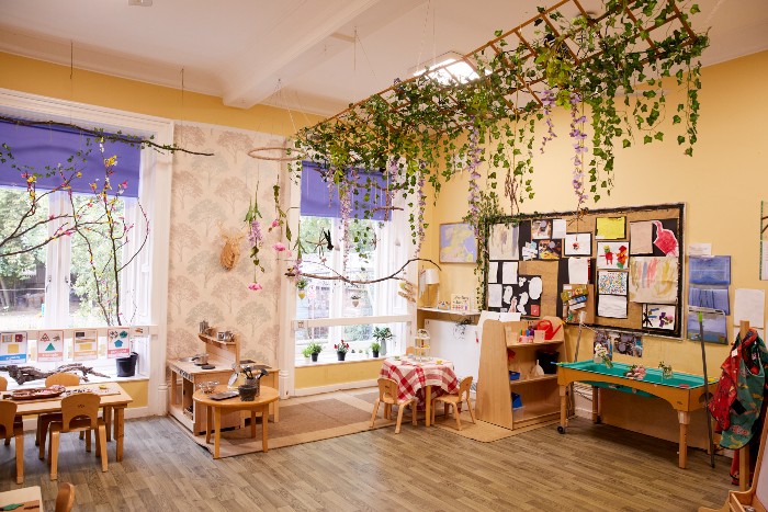 A windowed playroom in the nursery with small table and chairs positioned around the room. There are hanging plants on the ceiling above a small bookcase and sink