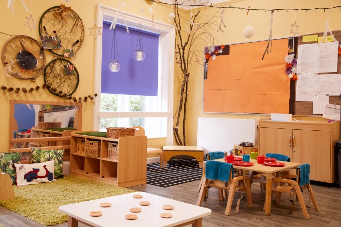 Children's playroom in the nursery. There is window in the back corner with dreamcatchers and a mirror next to it. There is a carpeted are in front of the mirror with cushions and storage cubbies next to it. There is a small table and chairs with plastic plates, cups and cutlery set out  