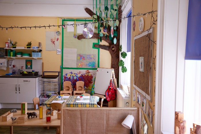 A playroom in the nursery with a whiteboard in the back corner and sink and cabinet area. There is a small table covered in a checked vinyl table cloth with chairs surrounding. There is another table with wooden animal toys on it and crafted tree coming out of the wall next to the window on the right 