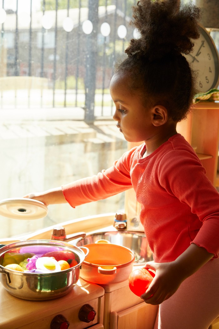 A young black girl next to a window playing with plastic kitchen utensils and food 