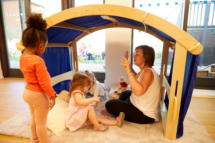 Two children play with a member of nursery staff under a covered play area indoors