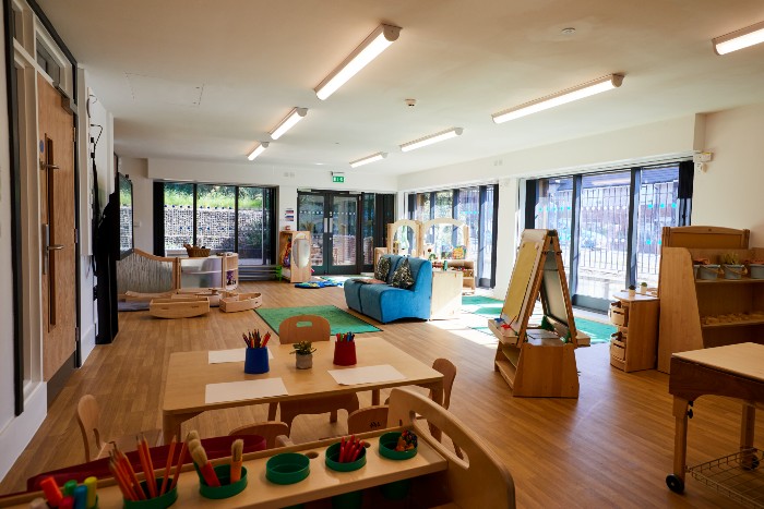 Interior of the Meadows nursery. Floor to ceiling windows on the back and right wall. A small seat with a carpeted area, play area and easel with paper. In the foreground there is a small table with paper and a cart filled with art supplies