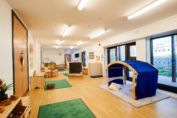Interior shot of the Meadows nursery. The room is long with a glass door and windows on the right wall closest to the camera. In front of the window is a play area covered by a wooden frame with blue material. There are wooden doors on the left wall with 2 green rugs in front. In the background there is a table and chairs, a chalk board, bookcase and a door on the back wall