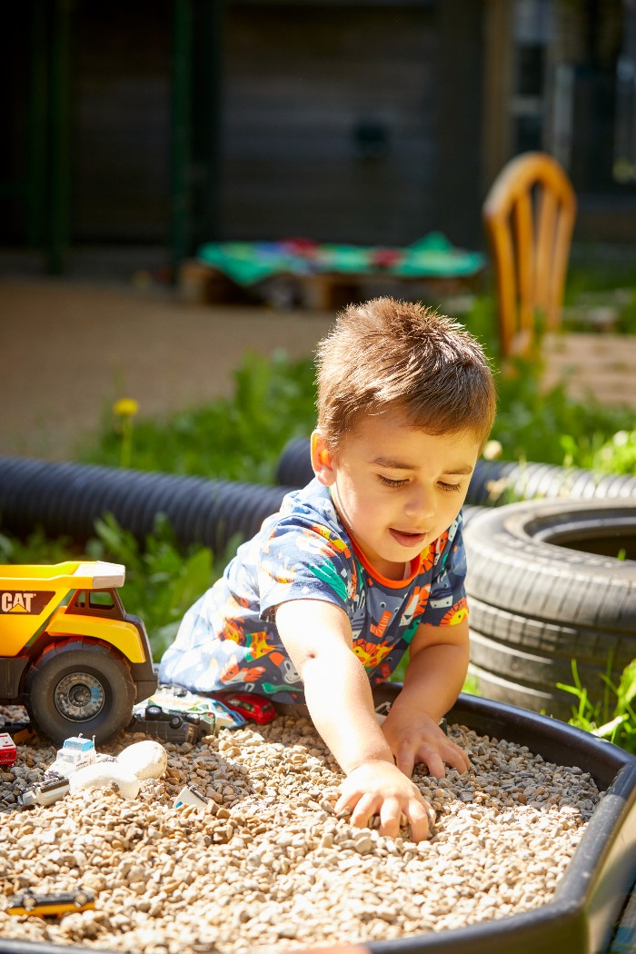 A young boy playing outdoors with a container filled with stones and various toy vehicles. There are repurposed tires, tube and table and chairs in the background