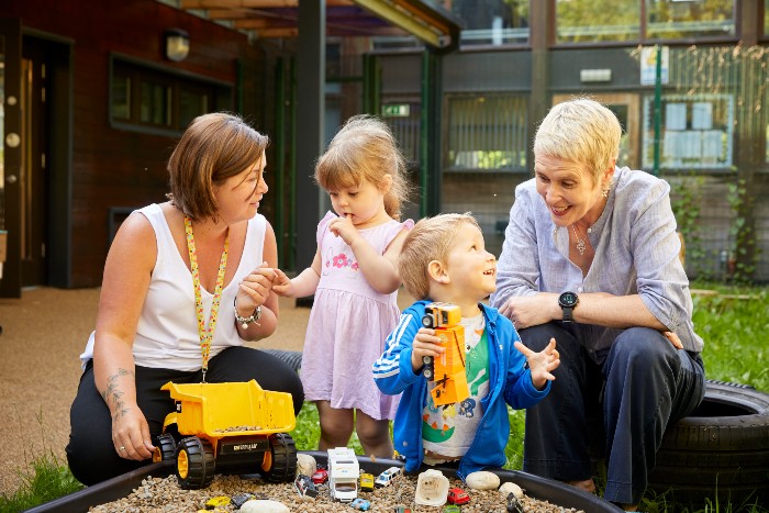 Two nursery staff and two young children playing outside in a sandpit filled with various toys