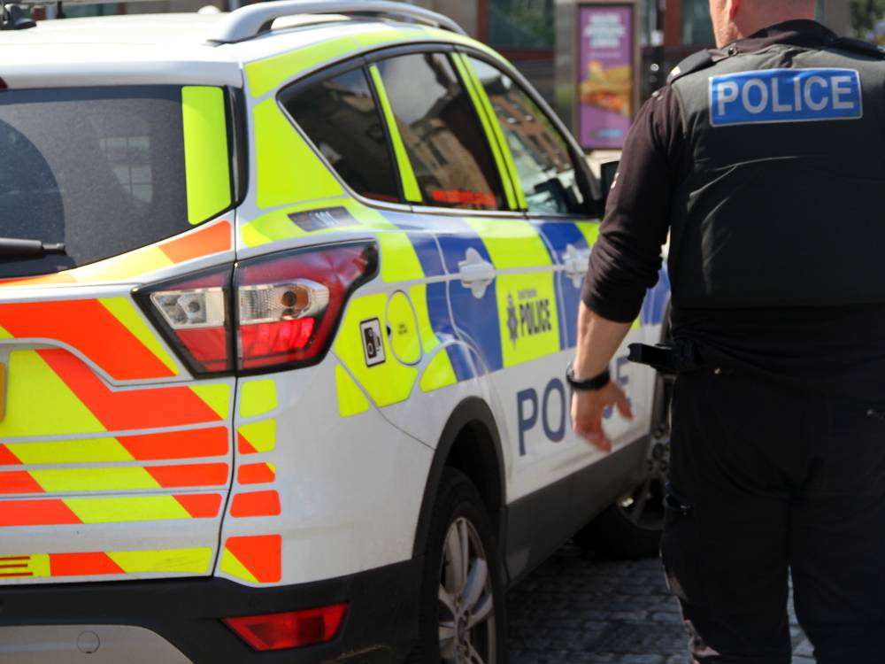 The rear side of a police car with reflective markings. A person wearing a police uniform and a vest labeled "POLICE" is standing next to the vehicle. 