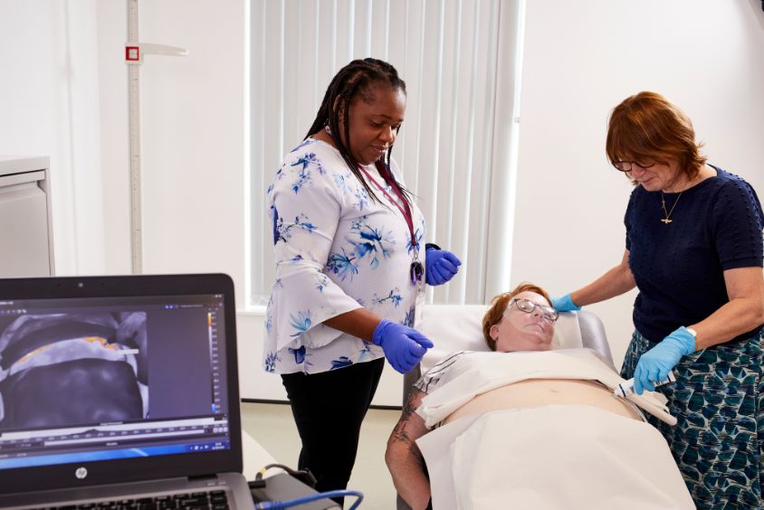 Two health and social care colleagues stand over a patient laid on a bed with an ultrasound device placed on their stomach within a clinical room, in the foreground a laptop is visible with the ultrasound imaging displayed.