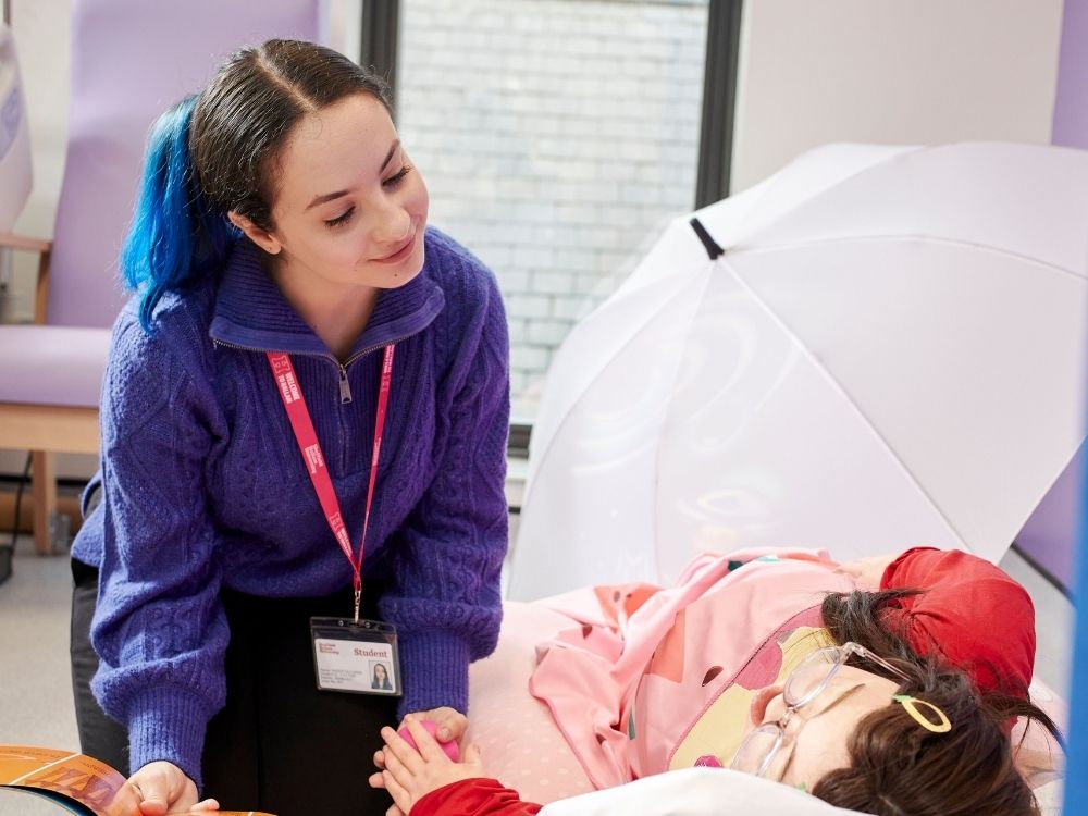 A student health visitor is wearing a blue jumper and red lanyard, interacting with a child manakin.