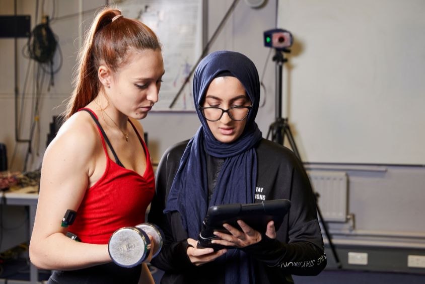 Two females stood side by side, one holding a weight in their arm whilst both look at the tablet screen held by the other.